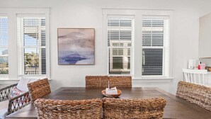 A dining area with a wooden table and wicker chairs, white walls, and large windows with blinds. - Sol Shine Gulf-View Retreat Steps from the Sand (Mexico Beach)