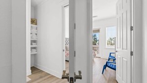 A hallway with white walls, a wooden floor, and a door leading to a room with a blue chair and a window.