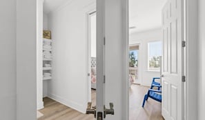 Un pasillo con paredes blancas, suelo de madera y una puerta que da a una habitación con una silla azul y un ventanal.