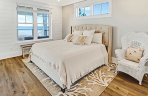 Un dormitorio con una cama grande, una silla blanca y vista al océano a través de una ventana.