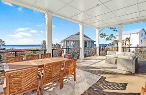 Un espacio al aire libre techado con una mesa y sillas de madera, un sofá de mimbre y vistas al océano y a casas de playa.
