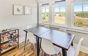 Un comedor con mesa de madera oscura y sillas blancas, ventanas amplias con vistas a un campo y una estantería con juegos de mesa.