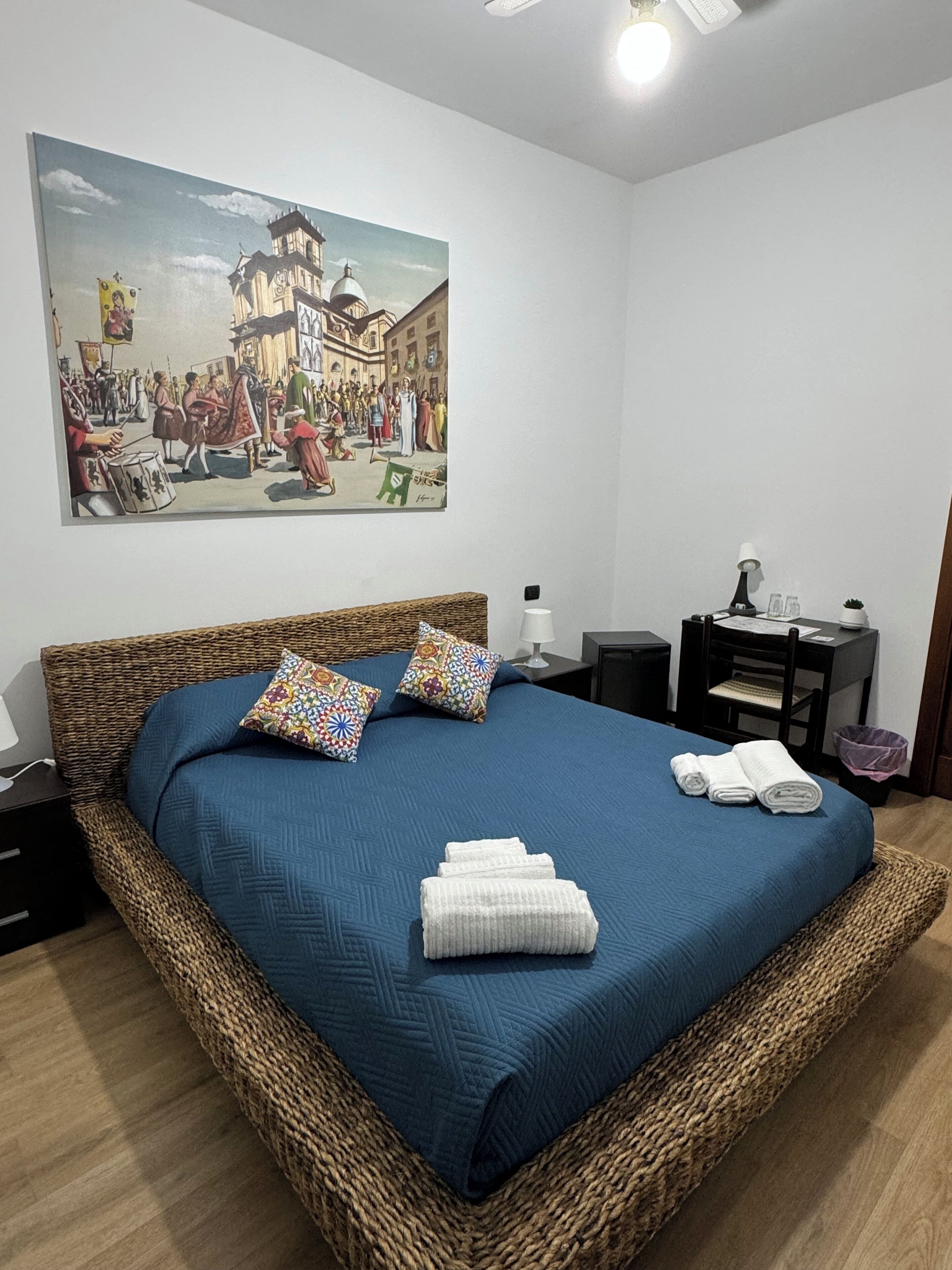 A bedroom with a large bed, a painting of a historical scene, a nightstand, a lamp, and a small table with a plant.