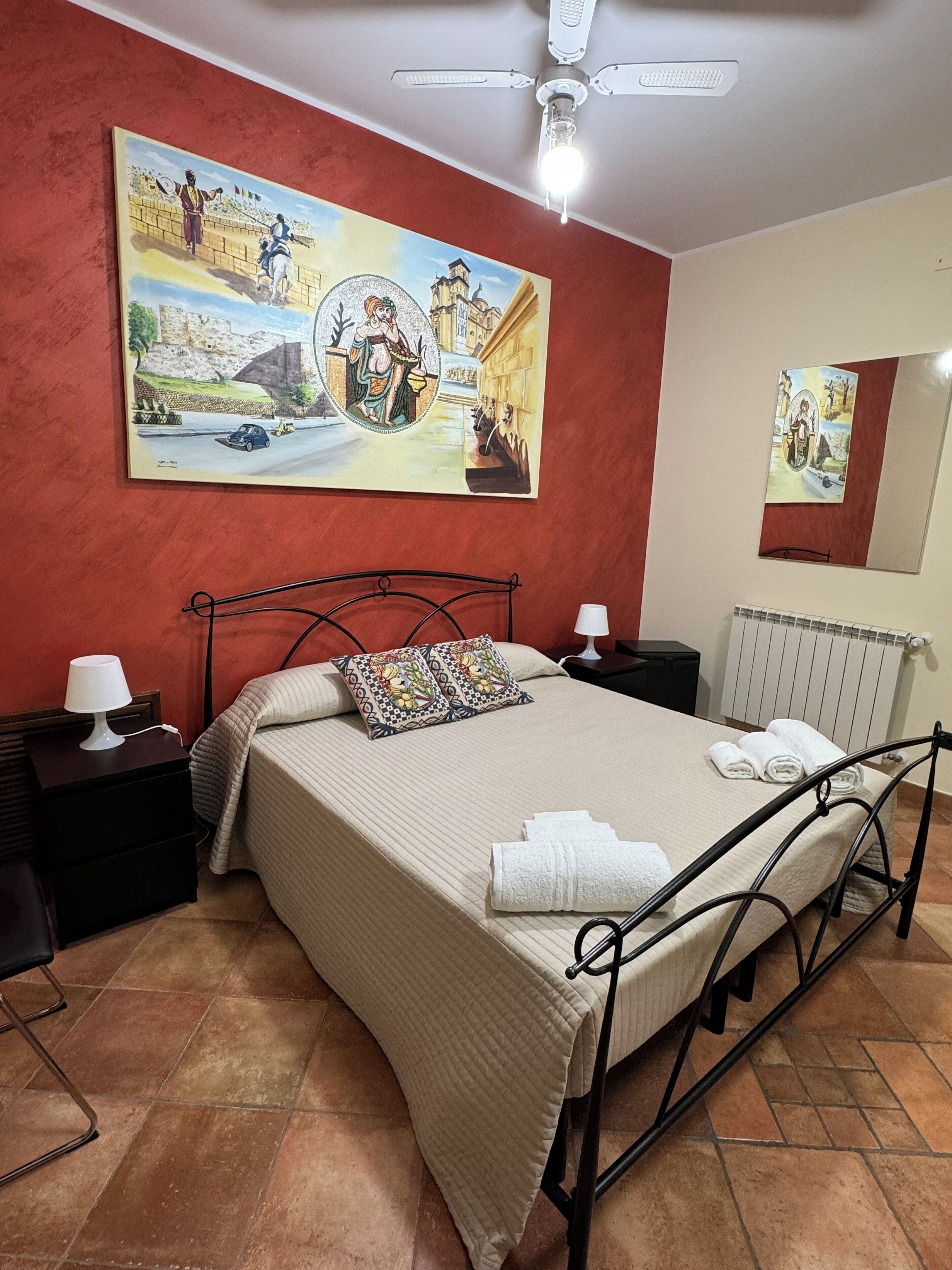 Un dormitorio con una cama, mesitas de noche, ventilador de techo y un cuadro grande en la pared.