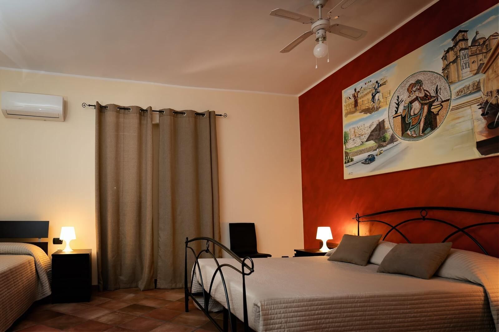 Une chambre d’hôtel comprenant un lit, deux tables de chevet avec des lampes, un ventilateur de plafond, une fenêtre avec des rideaux et un grand tableau mural.