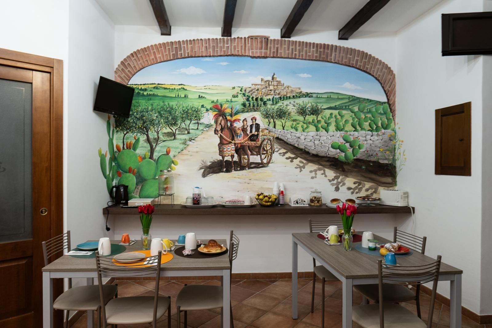 Un comedor con un mural de paisaje rural, un televisor montado en la pared y una mesa de buffet con diferentes alimentos.
