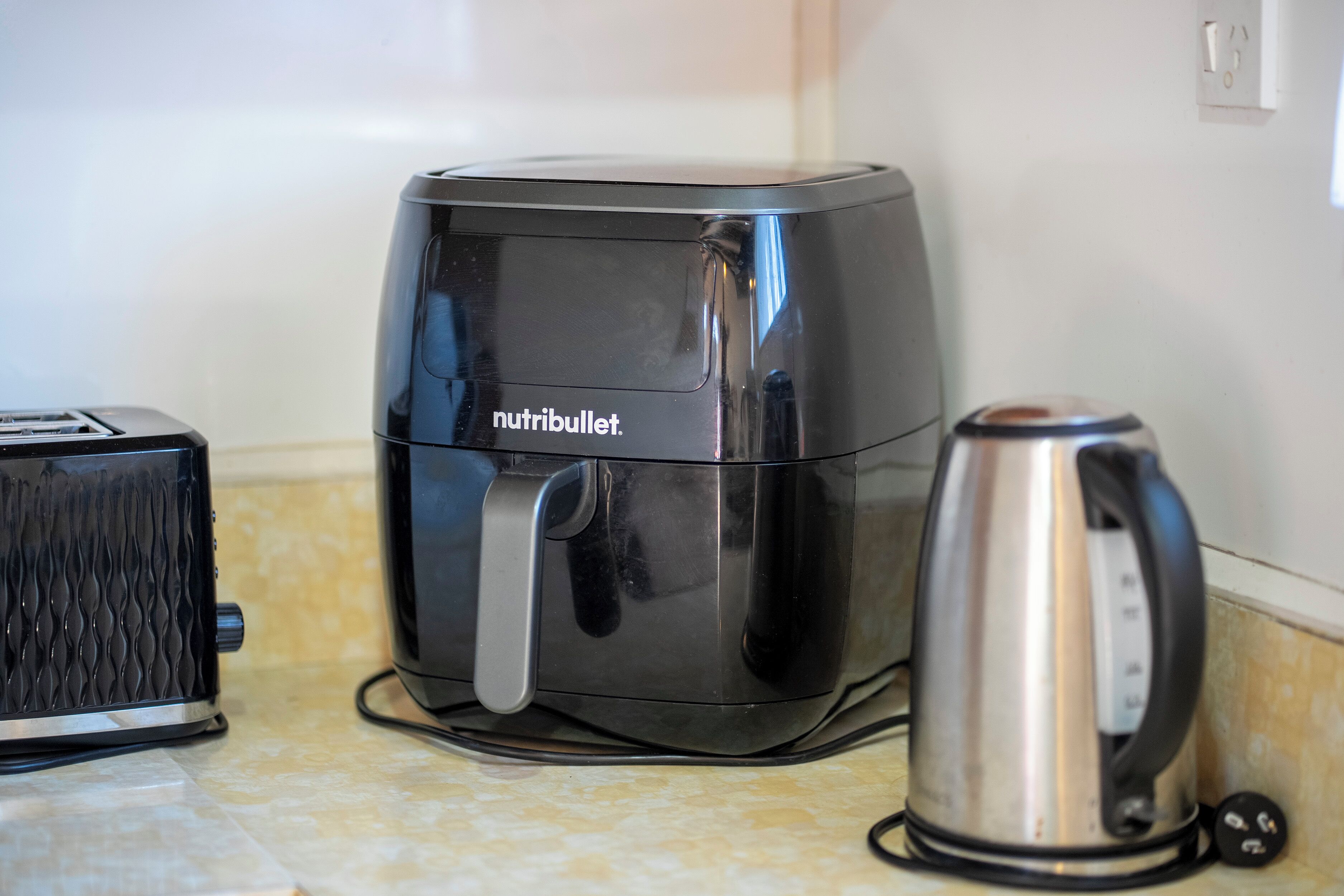 A kitchen with a black air fryer, a toaster, and a stainless steel kettle on a tiled countertop.