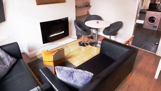 Una sala de estar moderna con un sofá negro, una mesa de centro de vidrio, un televisor y un comedor con una mesa pequeña y cuatro sillas.