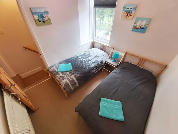 A small, cozy bedroom with two single beds, a window with a view of the outdoors, and a door leading to a staircase.