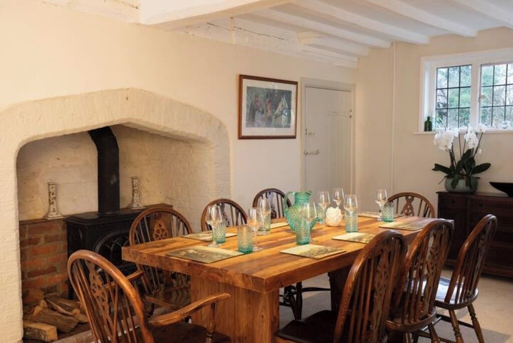 A dining room with a wooden table set for six, surrounded by wooden chairs. A fireplace is visible to the left, and a window with a view of greenery is on the right.