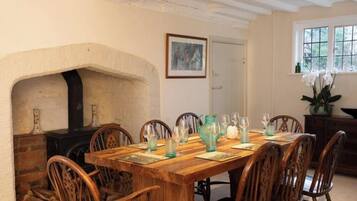 A dining room with a wooden table set for six, surrounded by wooden chairs. A fireplace is visible to the left, and a window with a view of greenery is on the right.