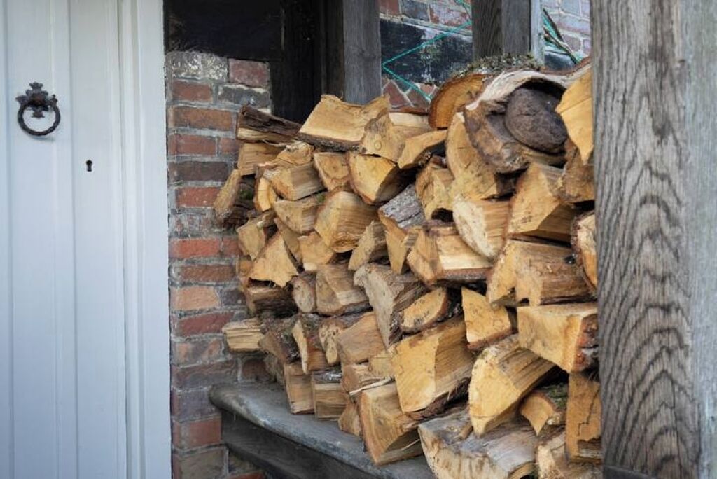 Une pile de bois de chauffage est appuyée contre un mur de briques, et une porte blanche se trouve à gauche.