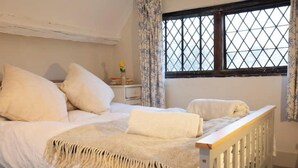 A bedroom with a bed, a nightstand, a window with a grid pattern, and a curtain. - Grade 2 Listed Rural Farm House (Chiddingfold)