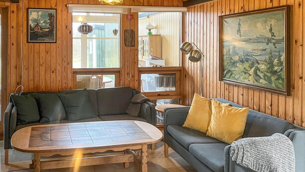 Ein gemütliches Wohnzimmer mit Holzwänden, einem grauen Sofa mit gelben Kissen, einem Holztisch und einem Landschaftsbild.