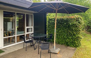 An outdoor patio with a table and chairs, a large umbrella, and a hedge in the background.