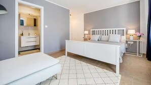 Une chambre à coucher moderne avec un lit blanc, une table de chevet avec une lampe, et une salle de bain visible à travers une porte ouverte.