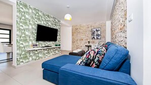 Un salon doté d’un téléviseur à écran plat fixé au mur, lequel est orné d’un motif de feuilles tropicales, d’un canapé bleu agrémenté d’un coussin à motifs floraux et d’un mur au revêtement imitant la pierre, situé derrière le téléviseur.