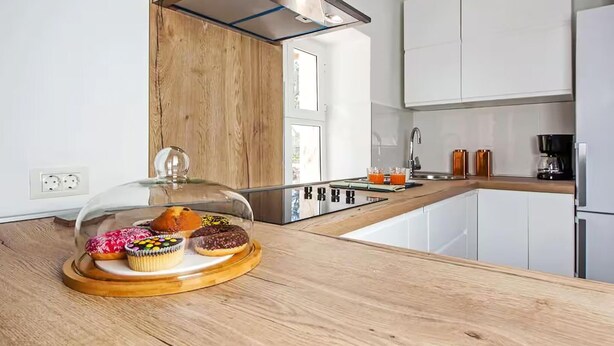 Une cuisine moderne avec des meubles blancs, un plan de travail en bois et une vitrine en verre où sont exposées des pâtisseries.