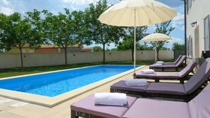 Un espace aménagé au bord de la piscine, avec des chaises longues et des parasols, une piscine à l’eau cristalline et une vue sur les arbres et les bâtiments en arrière-plan.