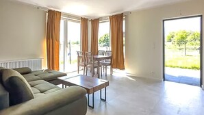 A spacious living room with a sofa, a coffee table, and a dining area with chairs and a table. There is a large window with curtains and a view of greenery outside. - 1 bedroom cozy apartment in Trzesacz (Trzesacz)