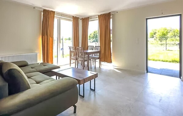 A spacious living room with a sofa, a coffee table, and a dining area with chairs and a table. There is a large window with curtains and a view of greenery outside.