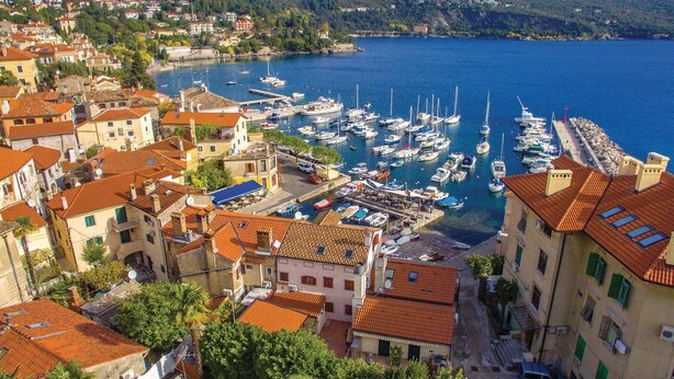 A coastal town with a marina filled with boats, traditional buildings with red-tiled roofs, and a hilly backdrop.