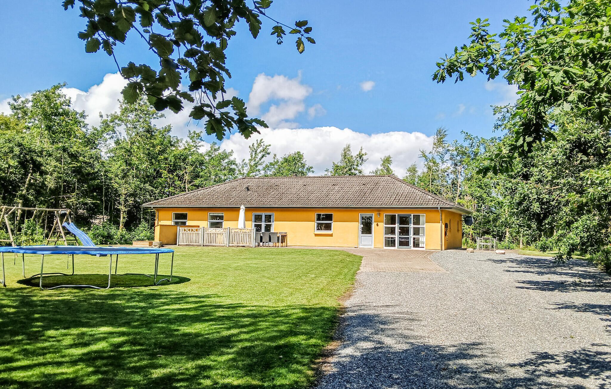 A yellow building with a tiled roof, a grassy yard, a trampoline, and a gravel path.