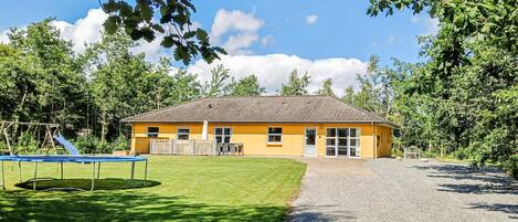 A yellow building with a tiled roof, a grassy yard, a trampoline, and a gravel path.