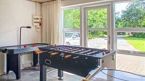 Une table de baby-foot dans une pièce dotée de grandes fenêtres, d’un radiateur fixé au mur et offrant une vue sur un parking extérieur.