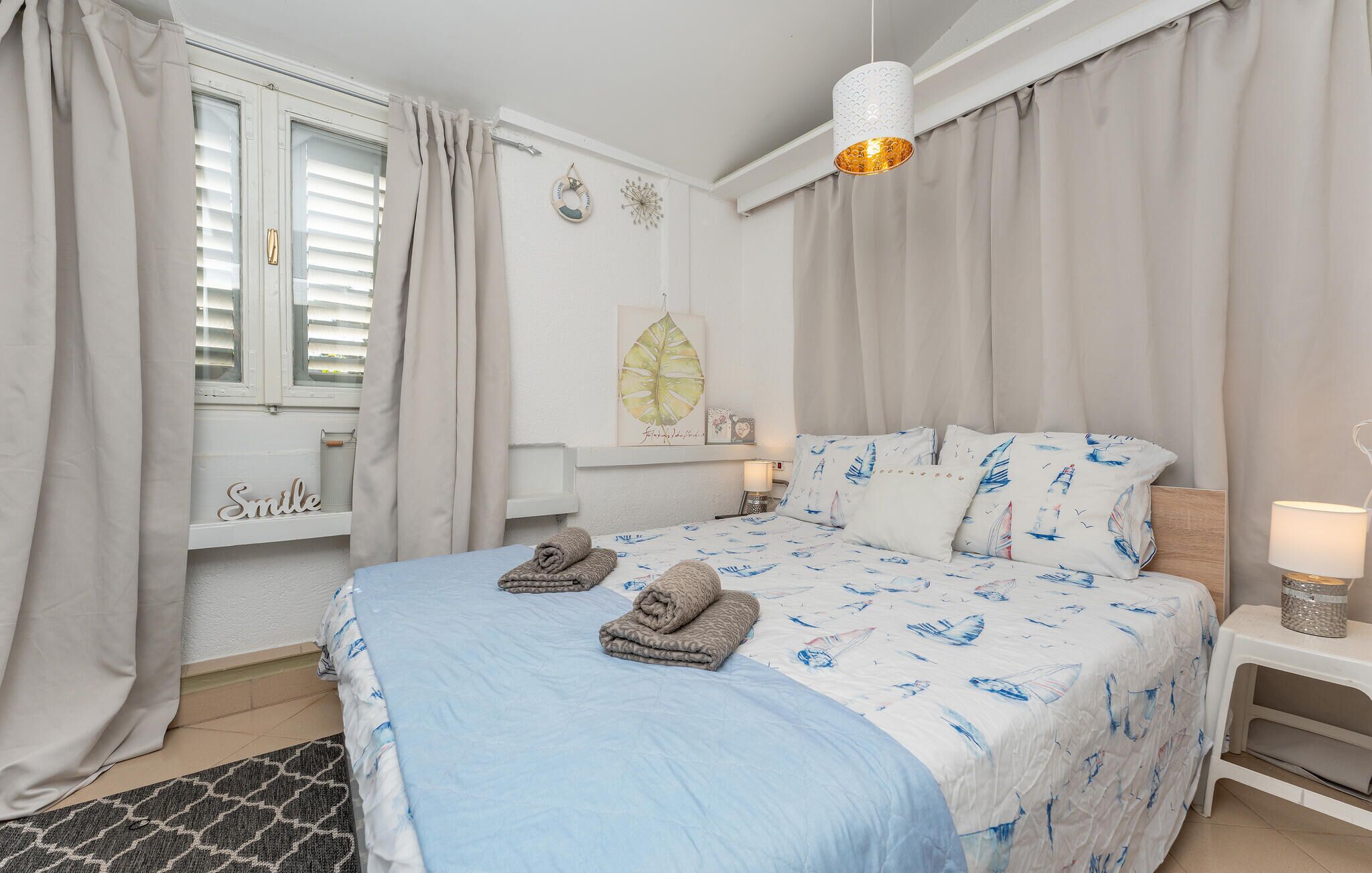 A bedroom with a bed, pillows, a bedside table, a lamp, a window with curtains, and a wall decoration.