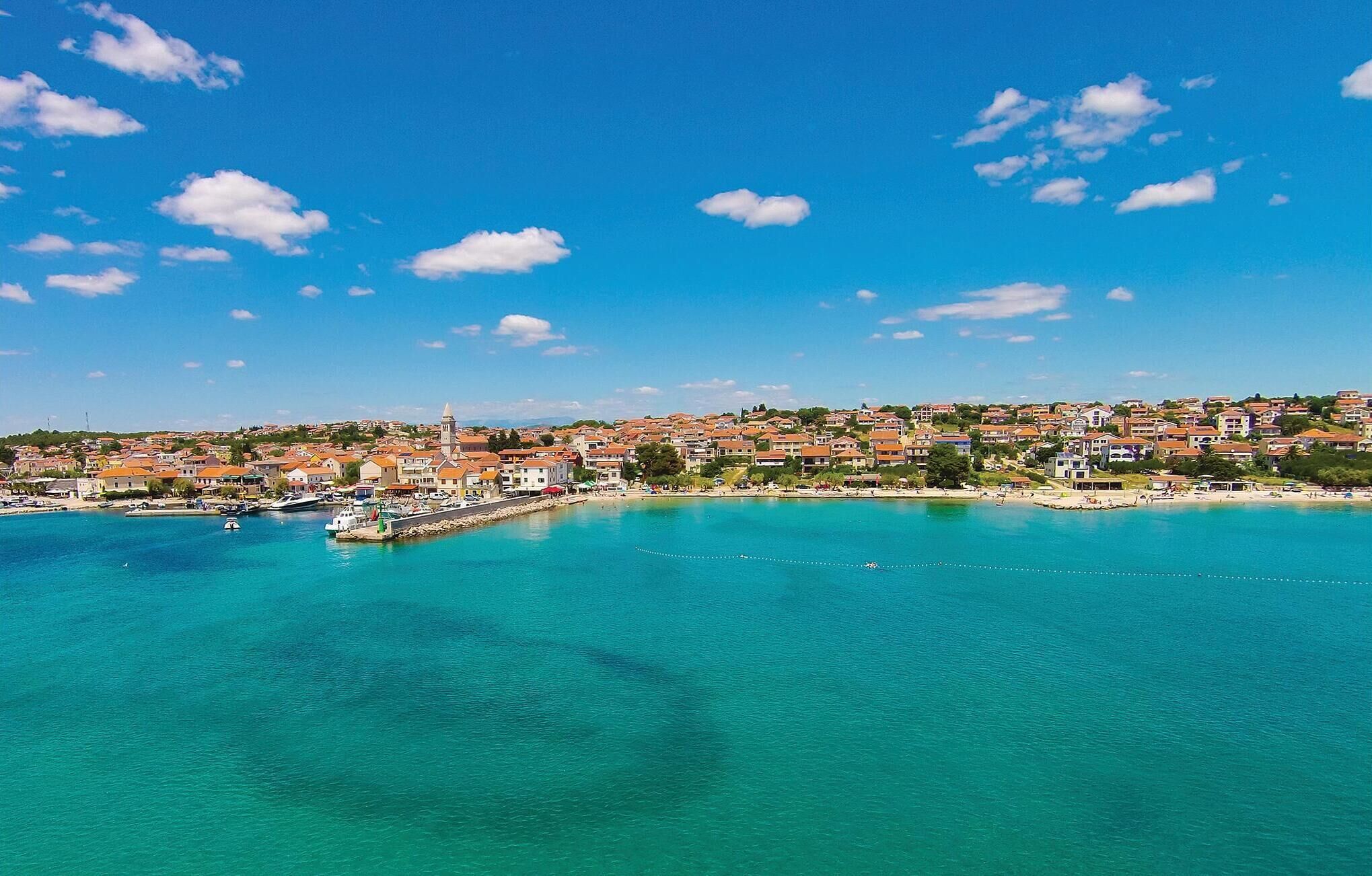 A coastal town with a clear blue sky, a harbor with boats, and buildings with red-tiled roofs.
