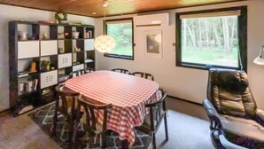 A dining area with a round table covered by a checkered tablecloth, surrounded by chairs. There is a bookshelf with various items on the left and a window with a view of trees on the right.