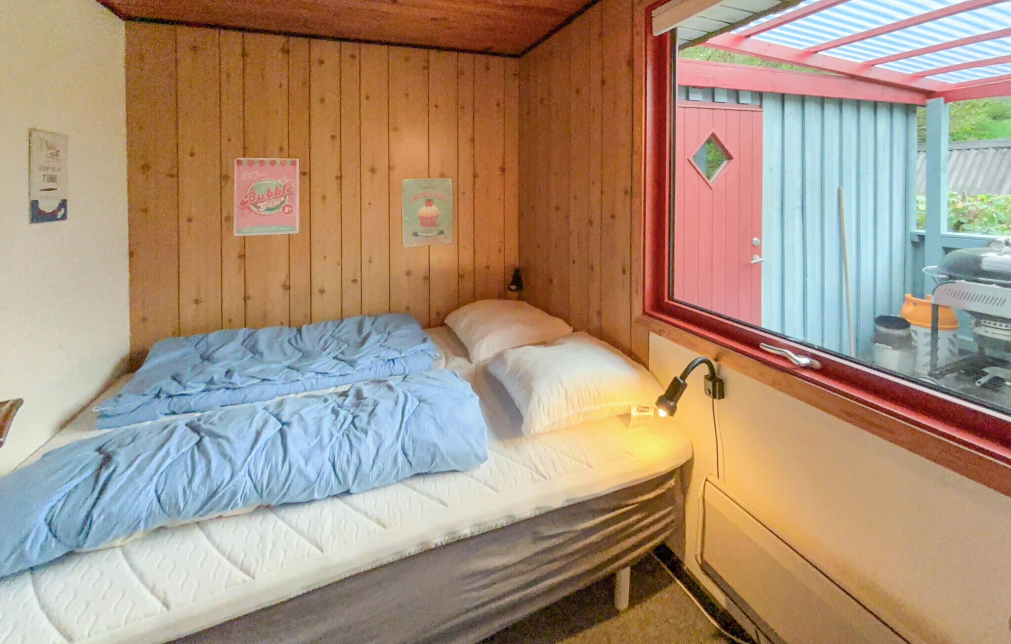 Een eenpersoonsbed met een blauwe deken en witte kussens in een kamer met houten wanden. Er is een raam met uitzicht op een rode en blauwe constructie buiten.