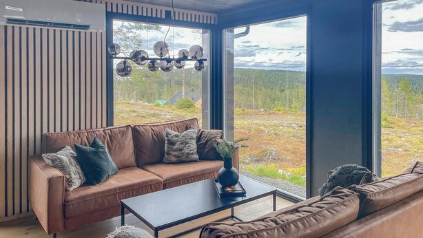 En moderne stue med en lædersofa i brunt, et sofabord og et stort vindue med udsigt over et skovlandskab.