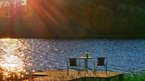 A wooden dock with two chairs and a table, overlooking a calm lake with reflections of trees and sunlight.