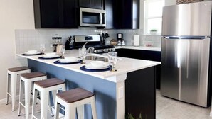 A modern kitchen with dark cabinets, a stainless steel refrigerator, a microwave, and a gas stove. There are bar stools and a small island with a sink and glassware. - Cozy Apartment in Austin by The Key Rentals (Austin)