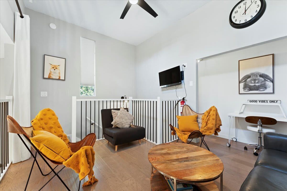 A modern living room with a black sofa, yellow chairs, a wooden coffee table, a TV mounted on the wall, and a clock.
