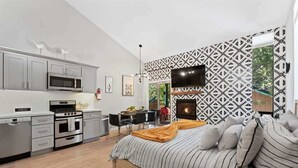 A modern kitchen with grey cabinets, a microwave, and a stove. There is a dining area with a table and chairs. A bed with striped bedding is in the foreground.