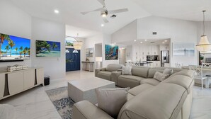 A spacious living room with a large beige sectional sofa, a flat-screen TV mounted on the wall, and a modern kitchen in the background.
