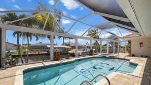 A swimming pool with clear blue water, surrounded by a covered patio area with palm trees and a view of a marina.