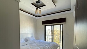 A bedroom with a large bed, a ceiling with recessed lighting, and a view of buildings outside through a window.