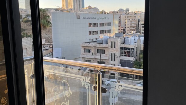A view from a balcony with a metal railing and decorative glass panels, overlooking a cityscape with buildings, including a bank with Arabic signage.