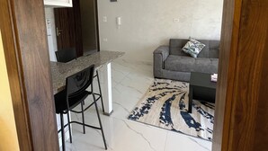 A modern kitchen with a marble countertop and black bar stools, a gray sofa with a patterned rug, and a wooden ceiling with exposed beams.