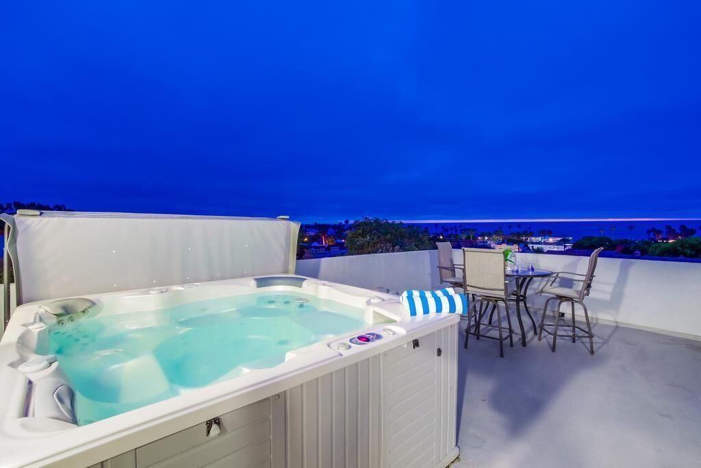 A hot tub on a rooftop terrace with a table and chairs, overlooking a cityscape at night.