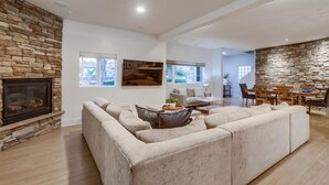 A spacious living room with a stone fireplace, a large beige sectional sofa, a dining area with a stone wall, and a view of the outdoors through large windows.