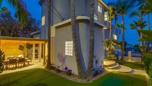 A modern two-story house with a covered patio, palm trees, and a well-maintained lawn.