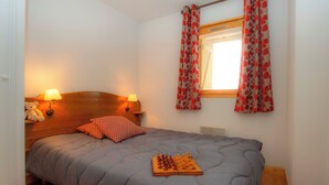 Un dormitorio con una cama, cabecera de madera, dos lámparas de noche, un osito de peluche, un tablero de ajedrez y una ventana con cortinas florales.