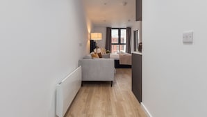 Ein schmaler Flur mit einem grauen Sofa und einer Lampe, der zu einem Raum mit einem Bett und einem Fenster führt.