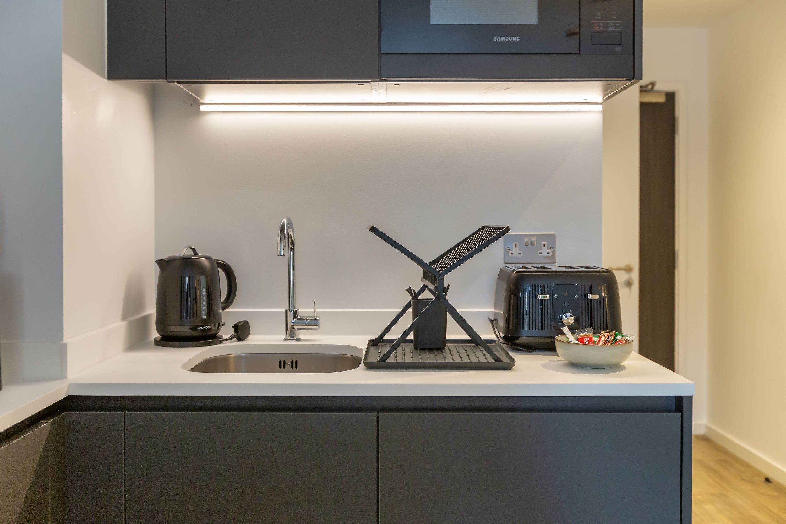 Une cuisine moderne équipée d’un four à micro-ondes encastré, d’une bouilloire, d’un grille-pain et d’un bol rempli d’en-cas posé sur le comptoir.