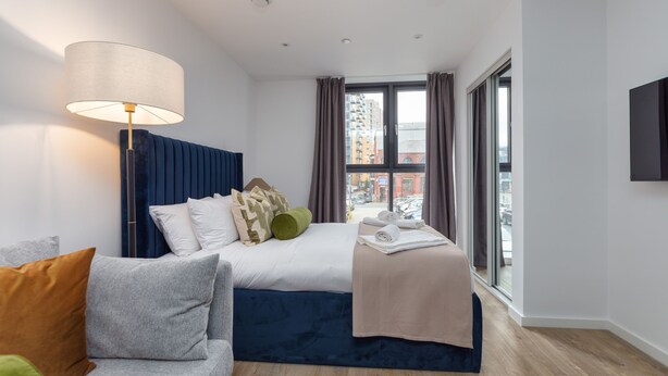 A modern hotel room with a bed, a sofa, a lamp, and a view of the city through large windows. - Chic New Cross Studio (Manchester)